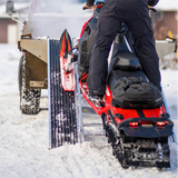 loading snowmobile on a caliber traction ladder in a snowy landscape with a vehicle in the background.